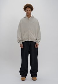 Felpa con zip beige, dotata di tasche frontali, vestibilità comoda e polsini a coste. Abbinata a jeans a gamba larga blu scuro e calzature color sabbia.