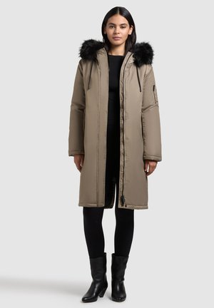 Long, beige padded coat with a fur-trimmed hood, featuring black accents, a zipper, and pockets. Paired with black leggings and boots.