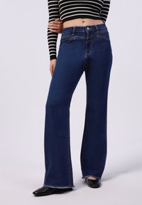 Donkerblauwe flare jeans met een hoge taille, voorzien van vijf zakken, lichte stiksels en rafelige zomen. Draag ze met zwarte puntige flats.