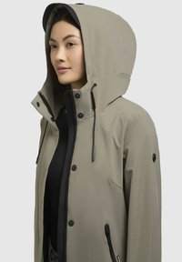 Giacca impermeabile beige con un ampio cappuccio, stringhe regolabili, chiusure a pressione e zip, e tasche laterali. Texture liscia e design minimalista.