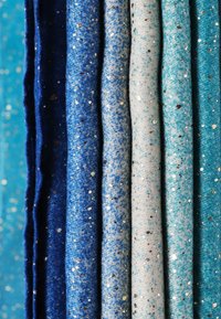 Cinque tessuti arrotolati in diverse sfumature di blu e acqua, decorati con piccole macchie d'argento e oro, presentano una superficie morbida e tessurizzata.