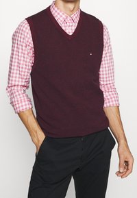 Bordeauxrode gebreide spencer met V-hals over een roze geruit overhemd. Zwarte broek, zichtbaar merkembleem op de spencer. Gladde textuur.