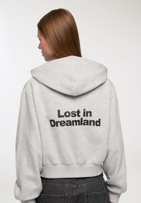 Szara, skrócona bluza z kapturem, z czarnym napisem "Zgubiony w Krainie Snów" z tyłu. Miękki materiał i dopasowane mankiety.