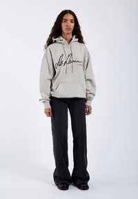 Sudadera gris claro con una gran impresión de firma negra, con un bolsillo canguro y capucha con cordón, combinada con vaqueros negros de pernera acampanada.