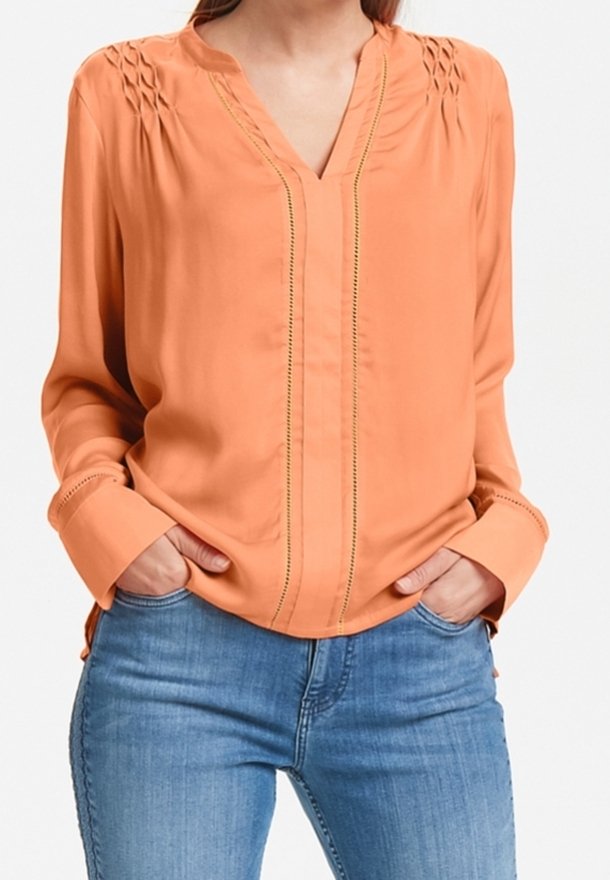 Blusa arancione con scollo a V, caratterizzata da dettagli arricciati, cuciture verticali e maniche lunghe. Abbinata a jeans azzurri chiari. Tessuto morbido.