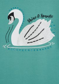 Tirkizna tkanina prikazuje grafiku bijele labudice s srebrnim detaljima, crnim obrubima i plavim draguljima, uz tekst "Sjaj i Sjaj".