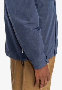 Marineblaue leichte Jacke mit einem Rautenmuster, langen Ärmeln und verstellbaren Bündchen. Verfügt über einen Kordelzug am Saum und ein Markenetikett am unteren Ende.