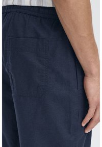 Mørkeblå bomuldsbukser med elastisk taljebånd, med en firkantet baglomme og et tekstureret stof. Nærbillede af sideudsigt.