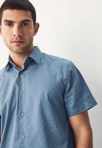 Chemise à manches courtes bleu clair avec un motif géométrique texturé, col boutonné et boutons nacrés. Finition du tissu lisse.