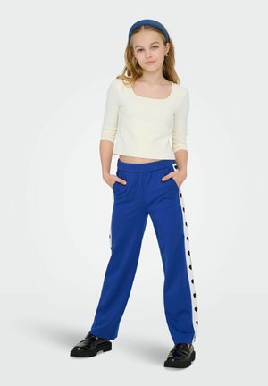 Blauwe wijde broek met zijknopen en zwarte hartpatronen, gecombineerd met een witte cropped top, gedragen met zwarte platform schoenen.