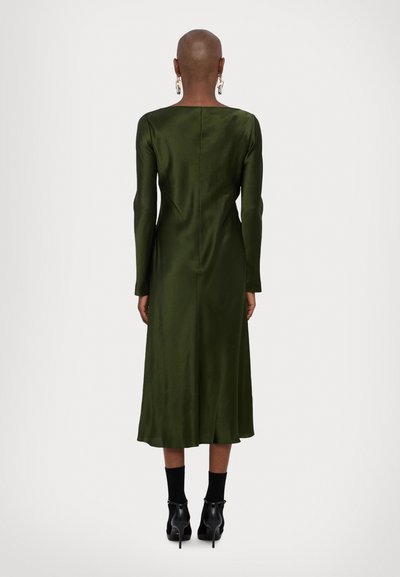 Donkergroen satijnen jurk met lange mouwen, rechte snit, naden aan de achterkant en midi-lengte, gecombineerd met zwarte enkelsokken en hakken.