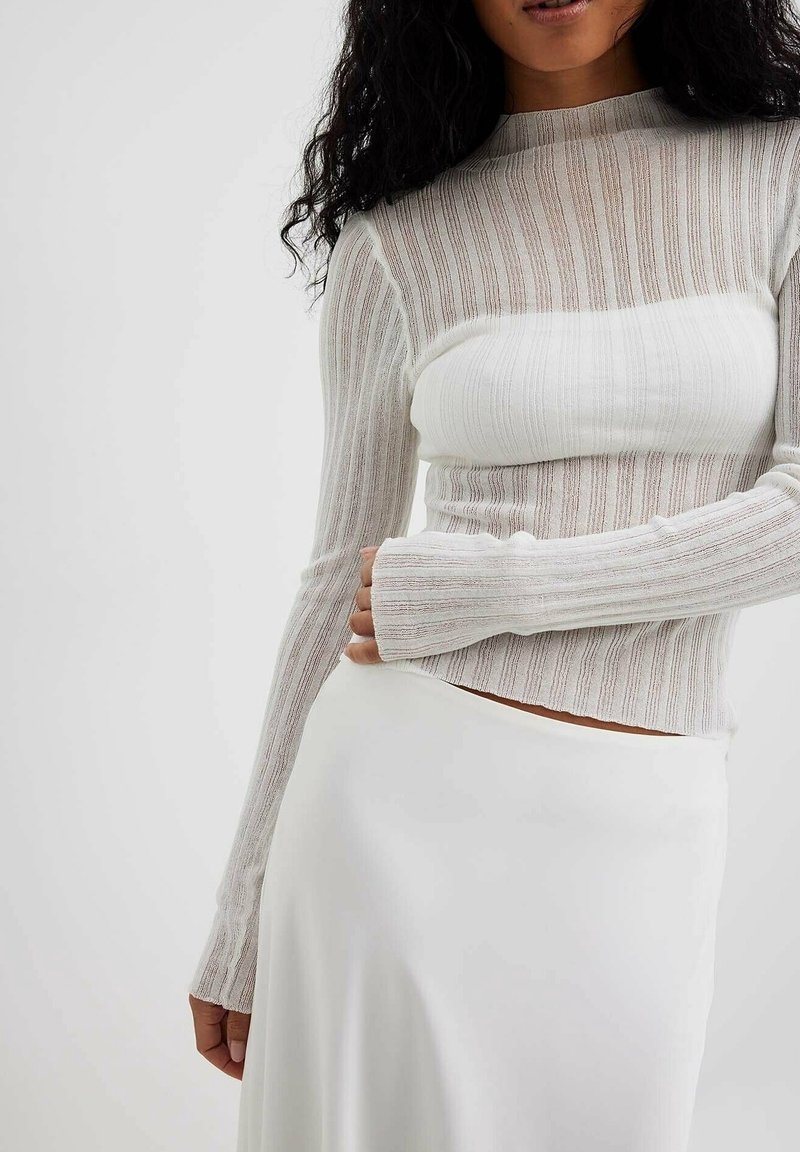 Femme portant un haut à manches longues, côtelé, semi-transparent beige et blanc, associé à une jupe blanche, sur un fond clair uni.
