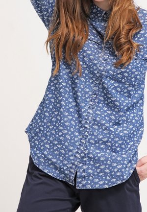Jong persoon met lang haar die een blauw bloemenoverhemd en donkere broek draagt, steekt een arm op tegen een effen achtergrond.