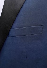 Tuvplāns ar tumši zila žaketa kabatu kreisajā pusē melnas atpogājamās atloka detaļas, redzama auduma faktūra un šuvju detaļas.