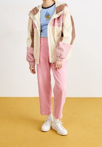 Persona con una chaqueta crema, rosa y marrón, un top corto azul con gráfico de rosa amarilla, pantalones rosa, calcetines blancos y zapatillas blancas de pie sobre un suelo amarillo.