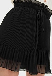 Falda negra plisada con tejido texturizado, cinturilla elástica y detalle de lazo; presenta un dobladillo con volantes y diseño en capas.