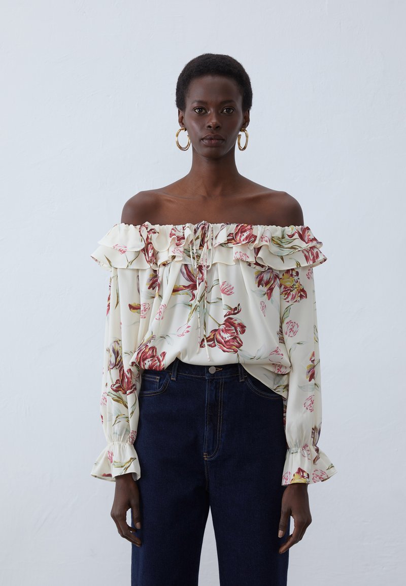 Blouse florale en off-the-shoulder avec des volants, fond crème et motifs de fleurs rouges et vertes. Associée à un jean en jean foncé taille haute.