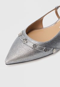 Zapato plano texturizado plateado con punta, que presenta una correa adornada con cinco pequeños adornos metálicos decorativos plateados.