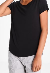 Svart kortärmad t-shirt med rund halsringning, mjukt tyg och en lätt rundad nederkant, bärs tillsammans med grå texturerade byxor.