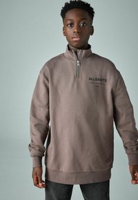 Felpa marrone con zip e collo alto, polsini a costine e dettagli del logo sulla parte anteriore. Texture morbida, vestibilità rilassata e design a strati.