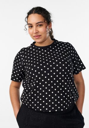 Kõrvarõngaid kandva lokkis juustega naine, kellel on seljas must lühikeste varrukatega särk valgete täppidega, käed taskutes, neutraalne taust.