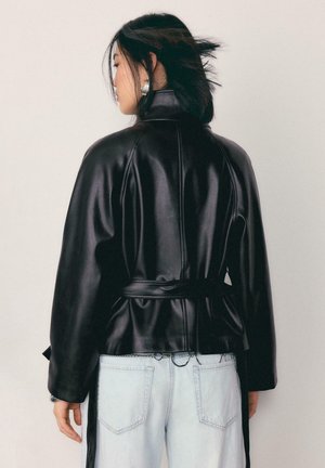 Kobieta o krótkich czarnych włosach, ubrana w czarną skórzaną kurtkę z paskiem i jasnoniebieskie dżinsy, stojąca bokiem na jednolitym tle.