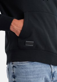 Felpa nera con tasca a marsupio, polsini a coste e una toppa con logo a contrasto. Il tessuto sembra morbido e flessibile, abbinato a jeans blu.
