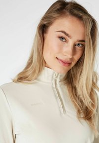 Valkoinen beige neljännes-vetoketjullinen pusero, joka on valmistettu pehmeästä kankaasta. Siinä on korkea kaulus, huomaamaton tuotemerkki-logo ja sileä pinta.