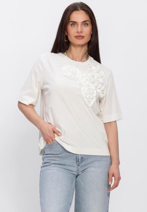 Femme aux longs cheveux noirs portant un haut blanc à manches courtes avec des détails floraux en 3D et un jean bleu clair, debout avec une main dans la poche.