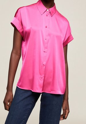 Femme portant un chemisier court en satin rose vif à manches courtes avec des boutons, associé à un jean bleu foncé, sur un fond neutre.