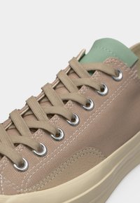 Μπεζ αθλητικό παπούτσι από καμβά με μπεζ κορδόνια, ασημένιες τρύπες, πράσινη υφασμάτινη γλώσσα και ελαστική σόλα εκρού.