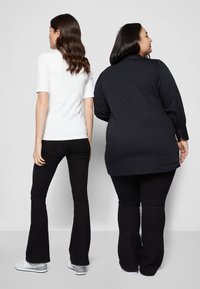 Duas pessoas estão de costas uma para a outra, uma vestindo uma blusa branca de mangas curtas com textura e jeans pretos de boca larga, e a outra com uma camisa preta abotoada e jeans.