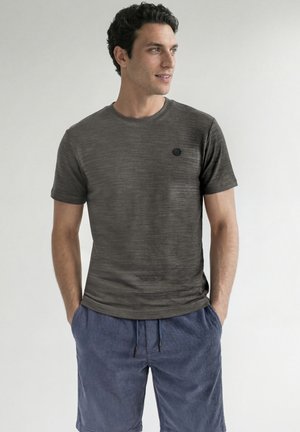 Mand iført en mørkegrå kortærmet t-shirt og blå kordshorts med hænderne i lommerne, kigger til siden mod en ensfarvet baggrund.