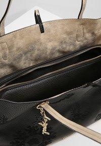 Käsilaukku mustasta karheasta nahasta, jossa on sileä kultasävyinen sisus, kaksinkertaiset vetoketjulliset osastot ja koristeellinen metallinen tarra hihnassa.