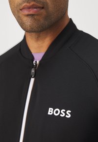 Svart zip-up jacka med hög krage, prydd med en vit "BOSS"-logotyp på framsidan och kontrasterande vita dragkedjedetaljer.