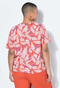 Persona con cabello corto que lleva grandes pendientes de aro, una camiseta de manga corta roja con patrones de hojas en rosa y púrpura, y pantalones naranjas con textura, de espaldas.