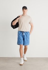 Chemise courte beige clair à manches courtes, shorts en denim bleu avec cordon de serrage et baskets blanches avec semelles beiges. Le modèle porte une veste noire.