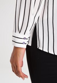 Blouse blanche à rayures verticales noires, tissu texturé, avec un poignet boutonné aux boutons violacés et une silhouette ample.