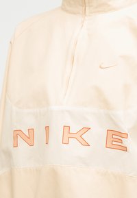 Μπεζ μπουφάν Nike με μισό φερμουάρ, πορτοκαλί περίγραμμα του κειμένου "NIKE" στο στήθος και μικρό πορτοκαλί λογότυπο Nike στην επάνω δεξιά πλευρά.