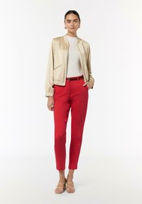 Hellbeige Bomberjacke mit elastischen Bündchen, getragen über einem weißen Top und roten Slim-Fit-Hosen. Deutliche Details sind ein Gürtel und Riemchensandalen.