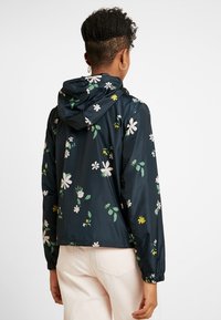 Niebieska kurtka przeciwdeszczowa w kwiaty z kapturem, elastycznymi mankietami i lekkim, błyszczącym materiałem w białe i żółte wzory kwiatowe.