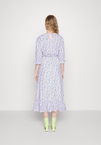 Žena s vlnitými blond vlasy má na sobě světle fialové květinové midi šaty s volánovým lemem a limetkově zelené kotníkové boty, stojí na jednobarevném pozadí.