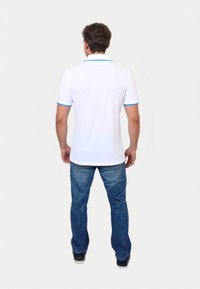 En ung mand står med ryggen til, iført en hvid polo t-shirt med blå kant og blå jeans, foran en ensartet lys baggrund.