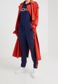 Un abrigo trench rojo sobre una camiseta navy con "KORS" en estampado metálico, combinado con joggers navy y zapatillas blancas.