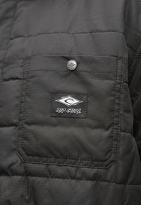 Czarna kurtka puchowa z przeszywanym kieszonką z srebrnym guzikiem i wyszywanym logo „Rip Curl”, prezentująca gładką, pikowaną fakturę.