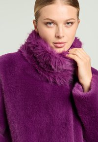 Abrigo de piel sintética púrpura con un cuello alto y texturizado. La tela parece suave y mullida, con un ajuste holgado y mangas anchas.