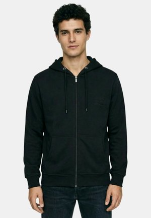 Jeune homme aux cheveux courts et bouclés, portant un sweat à capuche noir zippé avec poches avant et un jean foncé, debout devant un fond clair uni.