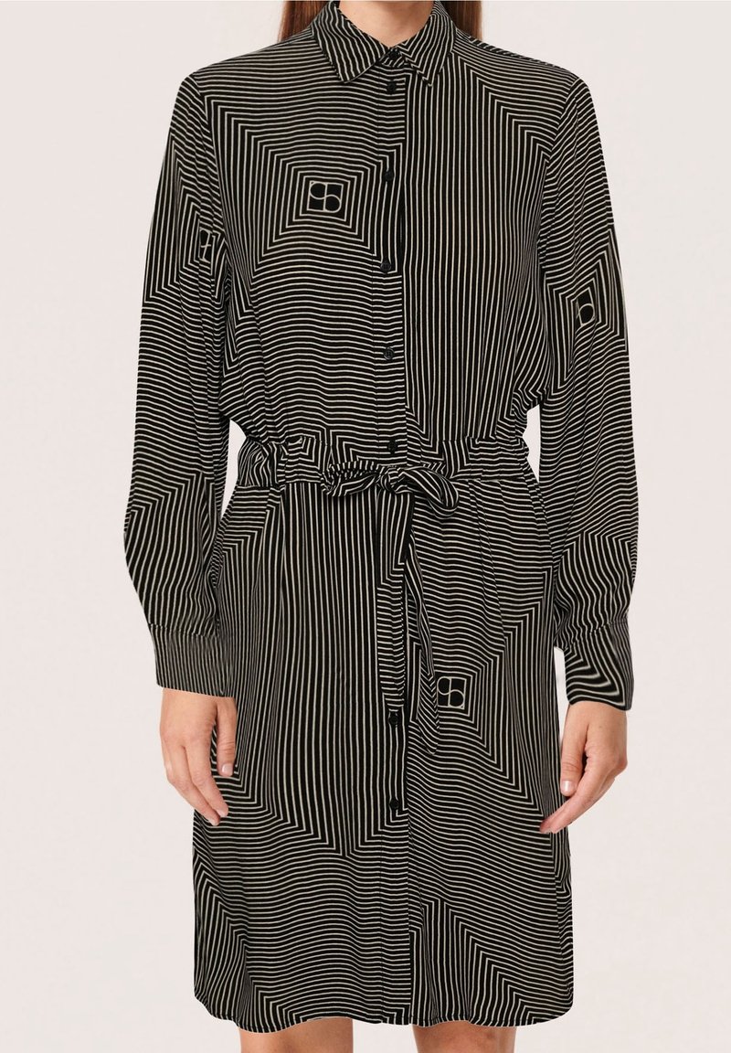 Vestito camicia a righe bianche e nere con colletto, maniche lunghe, chiusura con bottoni davanti, vita con cintura e motivi geometrici sul tessuto.