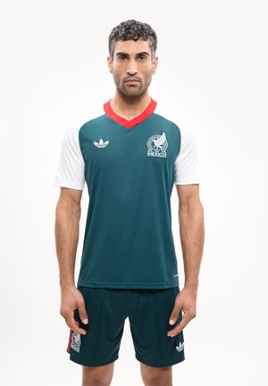 Άνδρας που στέκεται, φοράει φανέλα ποδοσφαίρου του Μεξικού με πράσινο σώμα, λευκά μανίκια, κόκκινο γιακά και αντίστοιχες πράσινες σόρτς με λογότυπα.