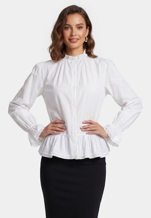 Vrouw met halflang bruin haar draagt een wit blouse met knopen, ruches aan kraag en manchetten, en een zwarte aansluitende rok, handen in de zij.